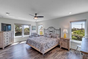 A bedroom with a large bed, a ceiling fan, a TV, a wooden dresser, and a window with a view of greenery. - Salt Therapy - Elevated house w/ beach access (St Augustine)