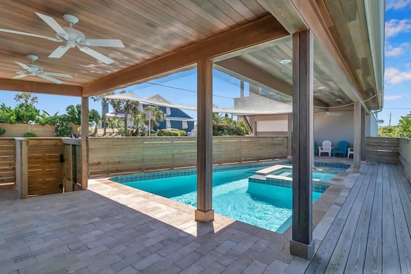 A covered outdoor swimming pool area with a wooden deck, ceiling fans, and a view of the surrounding neighborhood.