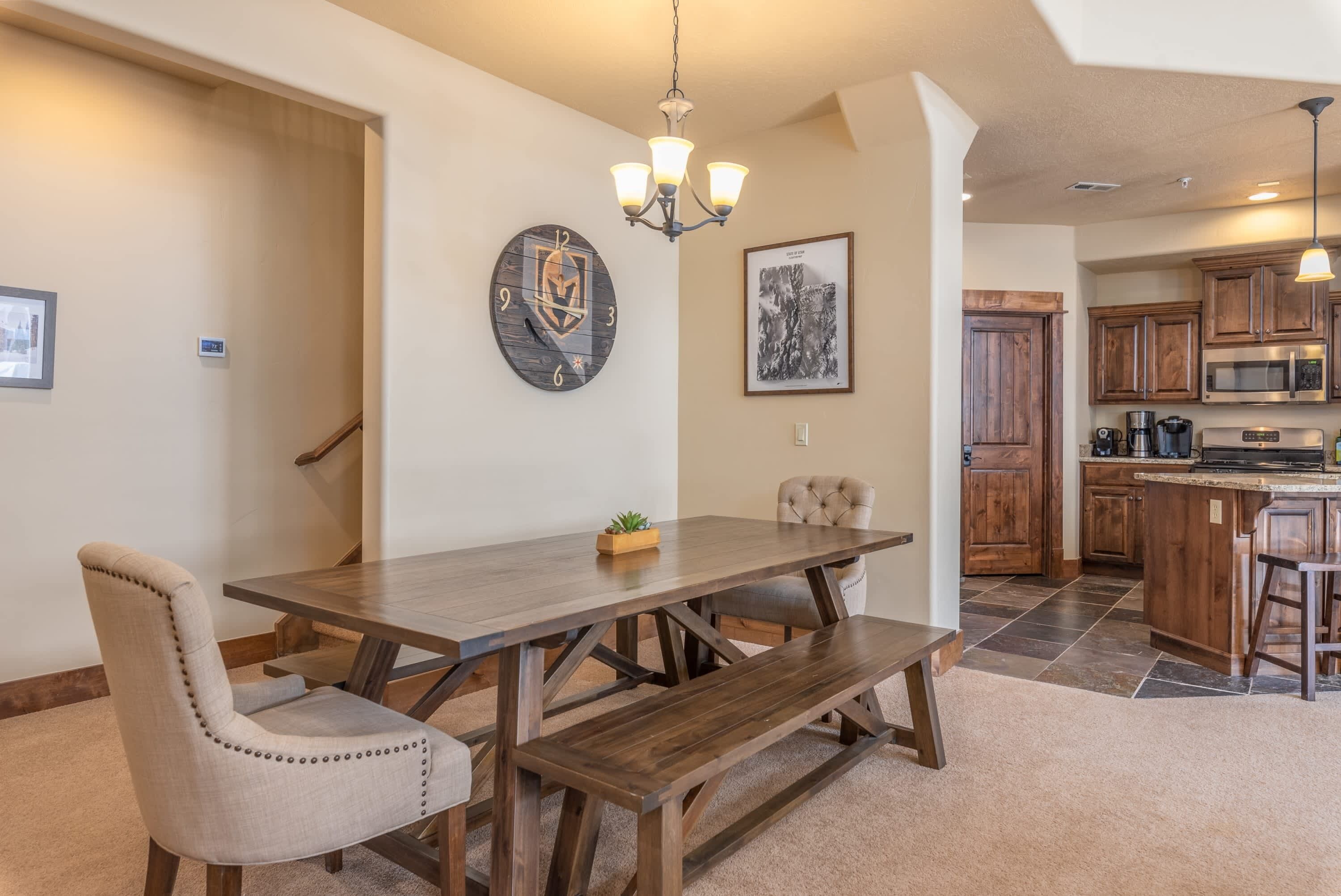 A dining area with a wooden table, cushioned chairs, a bench, a wall clock, a chandelier, and a kitchen in the background.