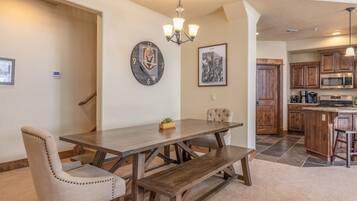 A dining area with a wooden table, cushioned chairs, a bench, a wall clock, a chandelier, and a kitchen in the background.