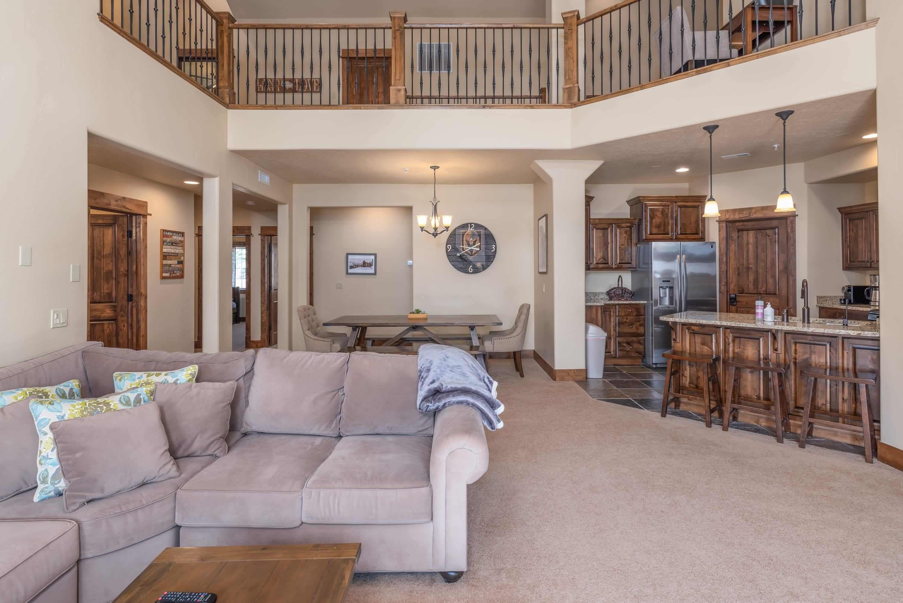 A spacious living room with a large beige sofa, a dining area with a table and chairs, a kitchen with wooden cabinets and stainless steel appliances, and a balcony with a wooden railing.