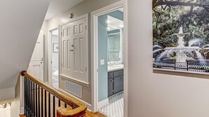 A hallway with a staircase, a bathroom with a glass shower, and a painting of a fountain. - Designer Furnished on Jones St, Heated pool access (Savannah)