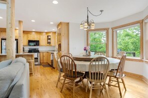A modern kitchen with wooden cabinets, a dining table with chairs, and a large window offering a view of greenery.