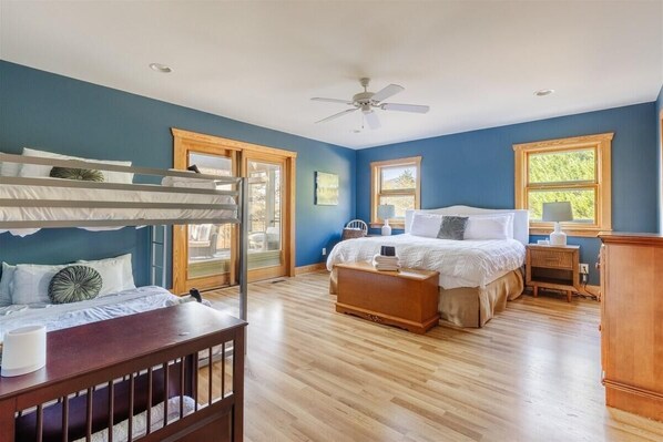 A bedroom with a bunk bed, a single bed, wooden flooring, a ceiling fan, and large windows.