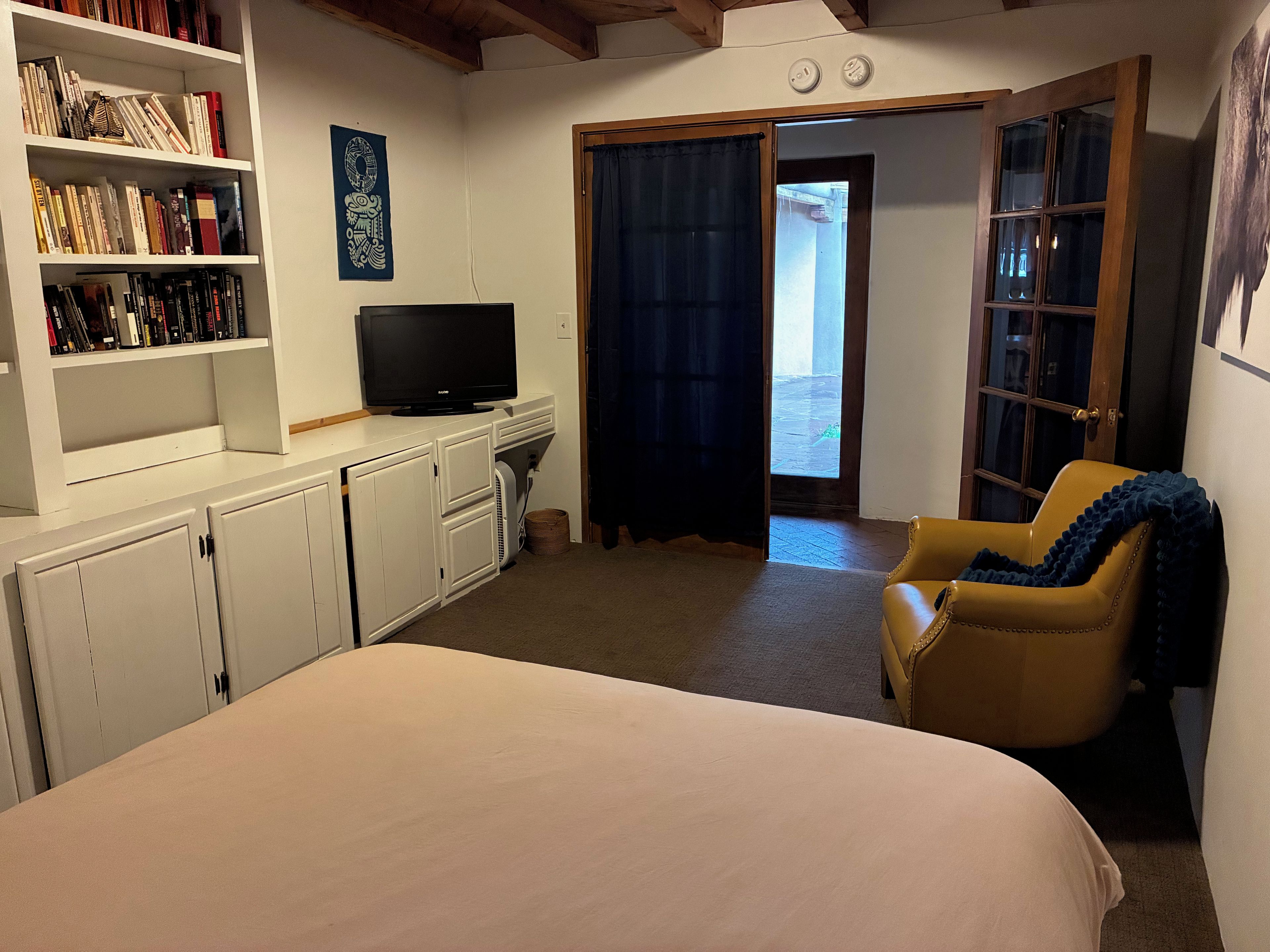 A hotel room with a bed, a television, a bookshelf, a chair, and a door leading to an outdoor area.