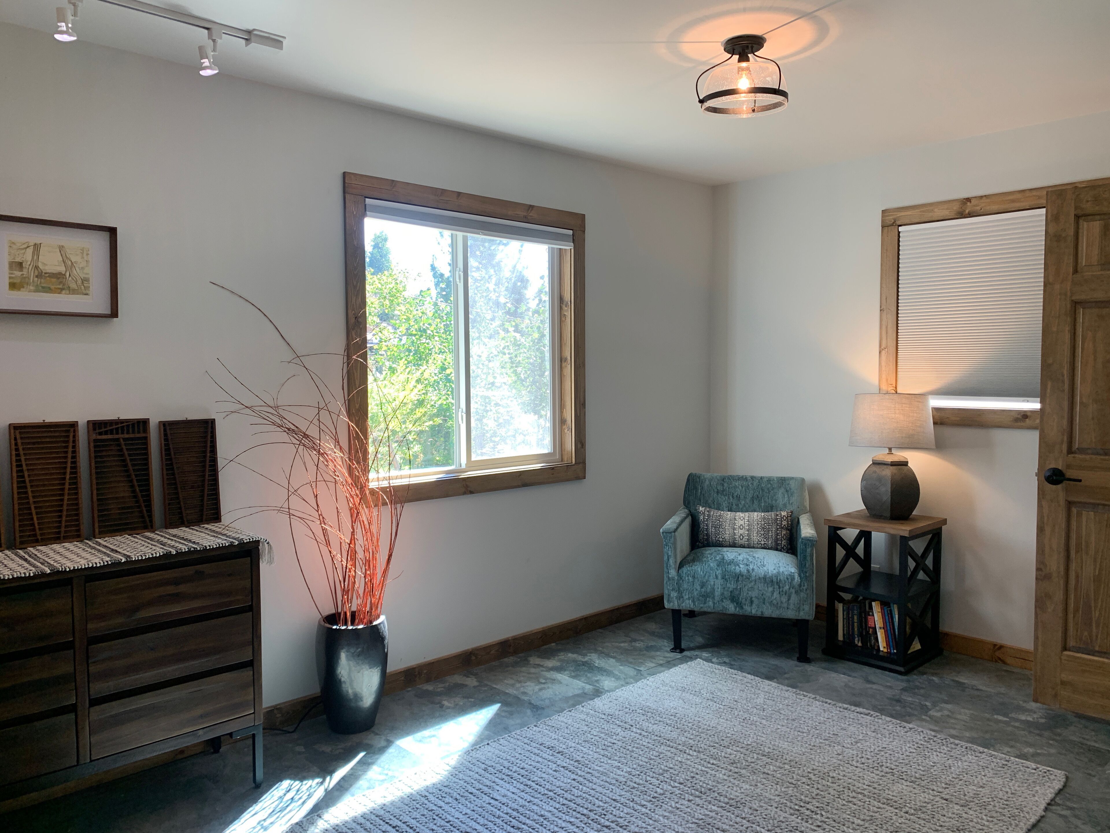 A room with a wooden dresser, a potted plant, a blue armchair, a small table with a lamp, and a window with blinds.