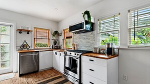 Cocina moderna con armarios blancos, electrodomésticos de acero inoxidable, encimeras de madera y ventanas amplias con persianas.