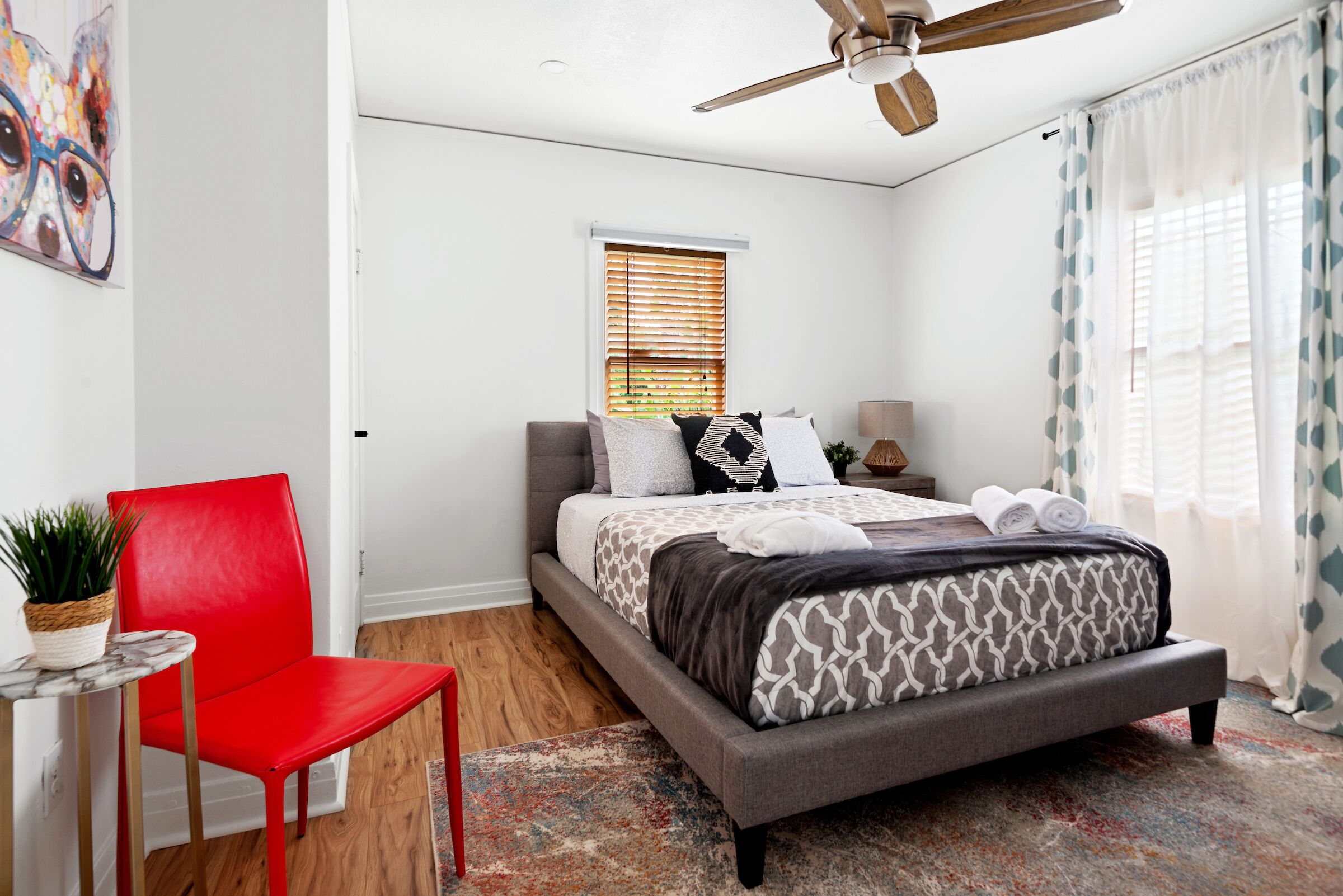 A bedroom with a bed, a red chair, a ceiling fan, a window with blinds, and a plant on a small table.