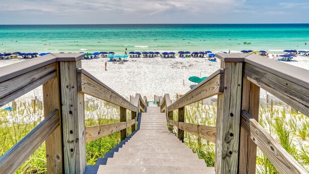 Eine Holztreppe führt zu einem Strand mit Sonnenschirmen und Menschen.