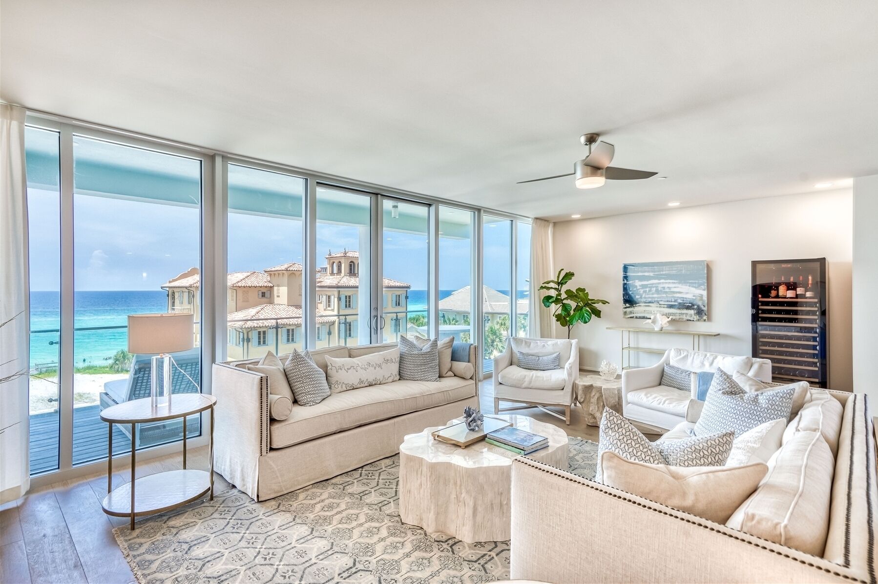 A spacious living room with large windows offering a view of the ocean and buildings, a ceiling fan, and a wine refrigerator.