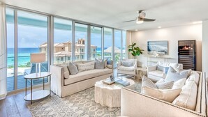 Ein geräumiges Wohnzimmer mit großen Fenstern, die Blick auf das Meer und Gebäude bieten, einem Deckenventilator und einem Weinkühlschrank.