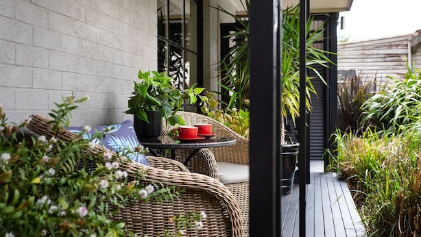 A wicker outdoor seating area with two red cups on a small round table, surrounded by lush greenery and a concrete wall. - East St 1 - tranquil escape, cosy charm (Daylesford)