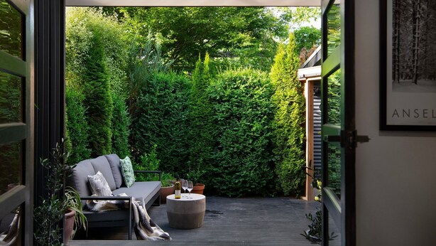 Een gezellig buitenverblijf met een bank en een ronde tafel, omringd door weelderig groen en een houten terras.