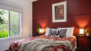Un dormitorio con un ventanal grande, una pared roja, una cama con ropa de cama estampada, dos mesitas de noche con lámparas y un cuadro enmarcado sobre la cama.