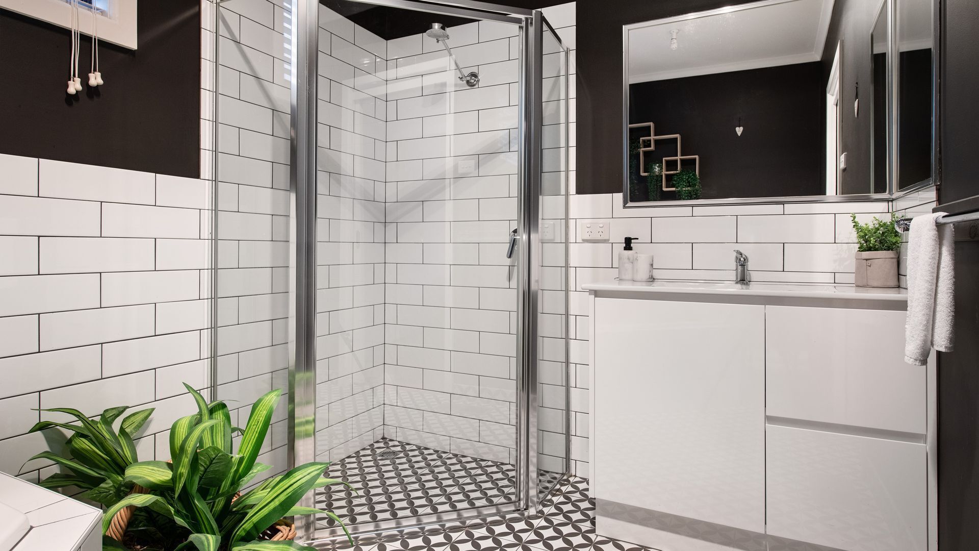 Une salle de bain moderne avec une douche à parois de verre, des carreaux de métro blancs, un sol à motifs géométriques, un miroir avec un cadre décoratif et une plante en pot.