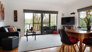 Ein modernes Wohnzimmer mit einer schwarzen Ledersofa, einem Holzesstisch mit Stühlen, einem Flachbildfernseher und einer großen Schiebeglasdoor zu einer Terrass.'