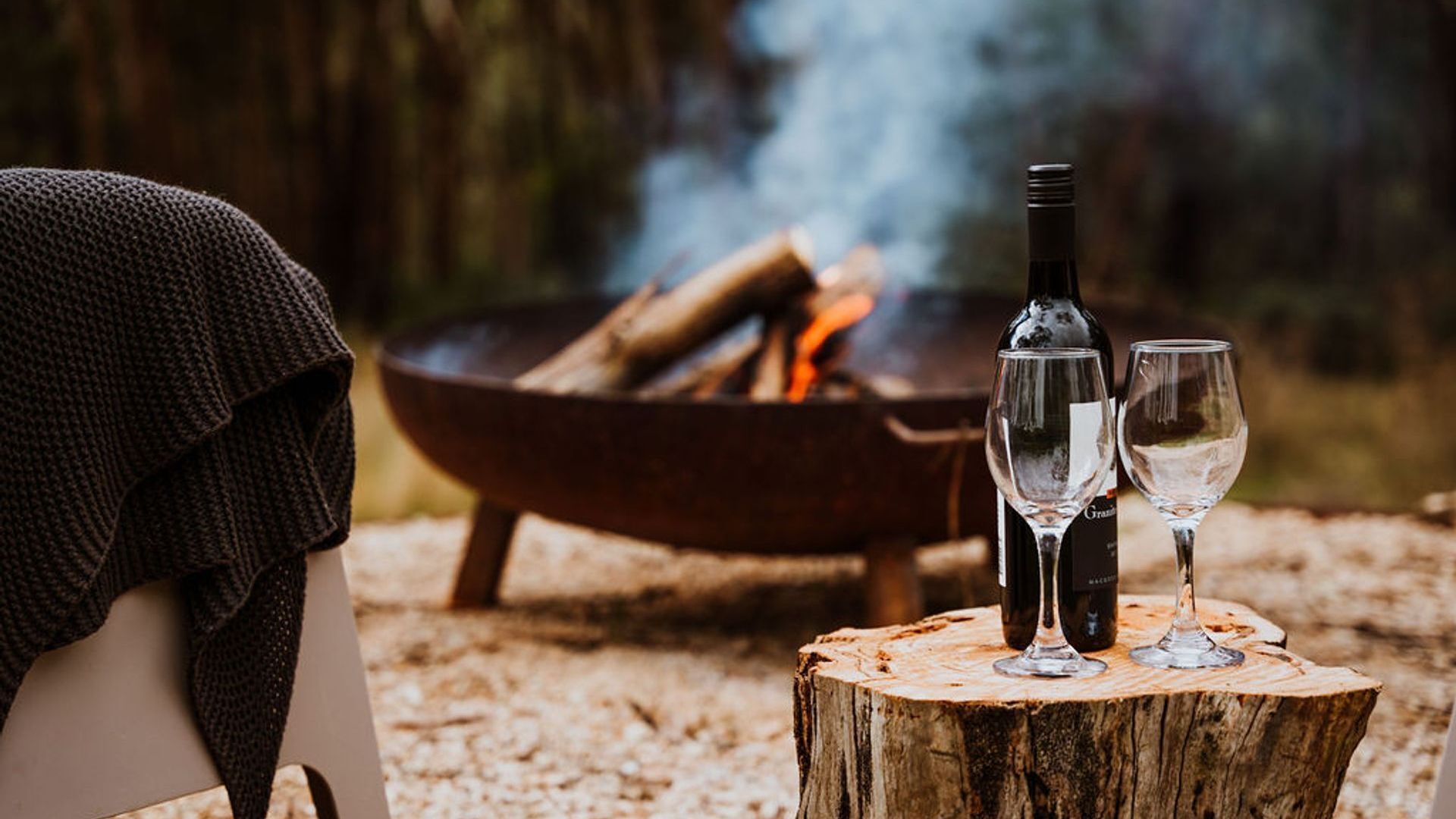 A cozy outdoor setting with a fire pit, two glasses, and a bottle, suggesting a relaxed outdoor dining experience.