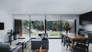 Un salon moderne doté d’une grande porte-fenêtre coulissante donnant sur un jardin. On y trouve un canapé, une table à manger avec des chaises, et un coin cuisine visible à travers la vitre.