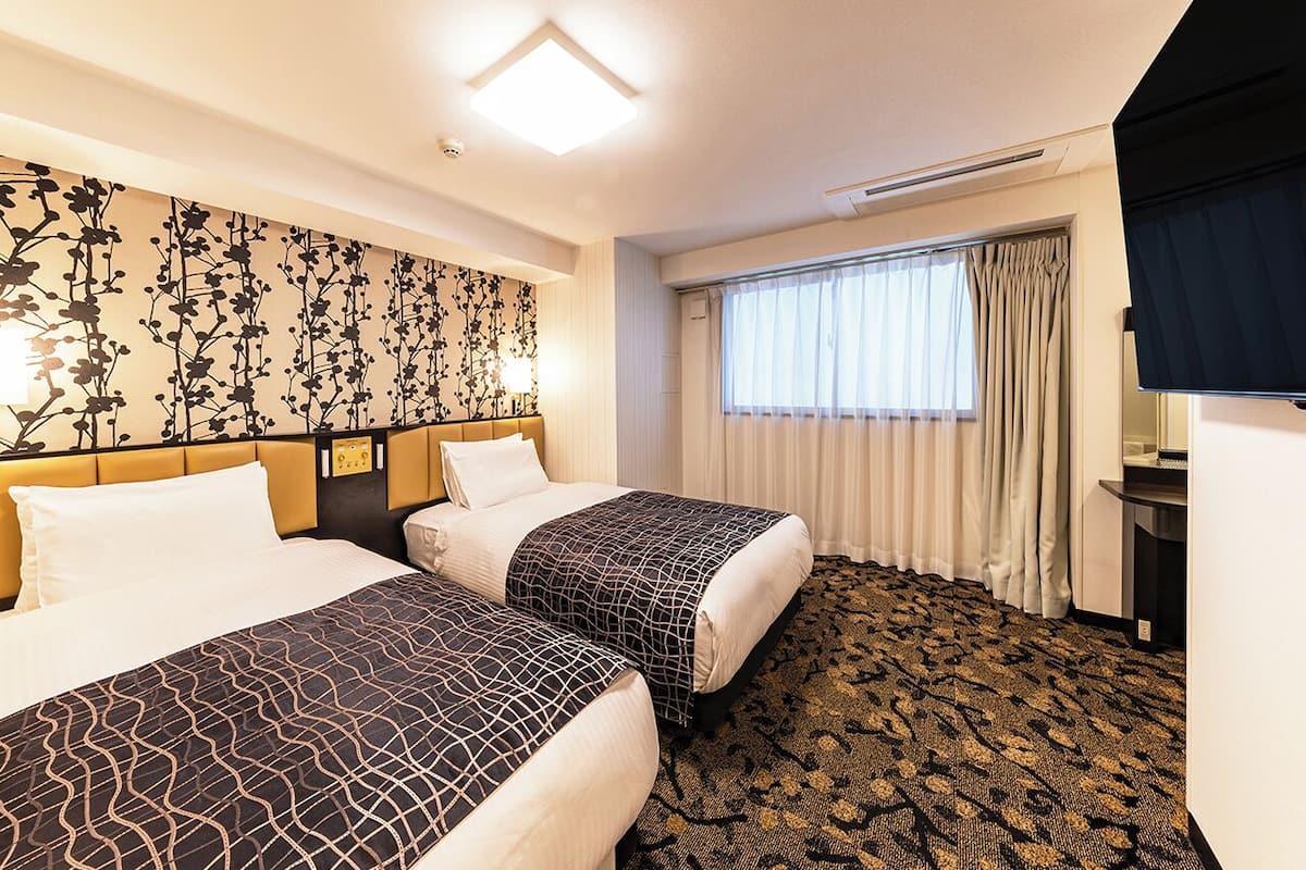 Ein Hotelzimmer mit zwei Betten, einem gemusterten Teppich, einem großen Fenster mit Vorhängen und einer Wand mit floralem Dekor.