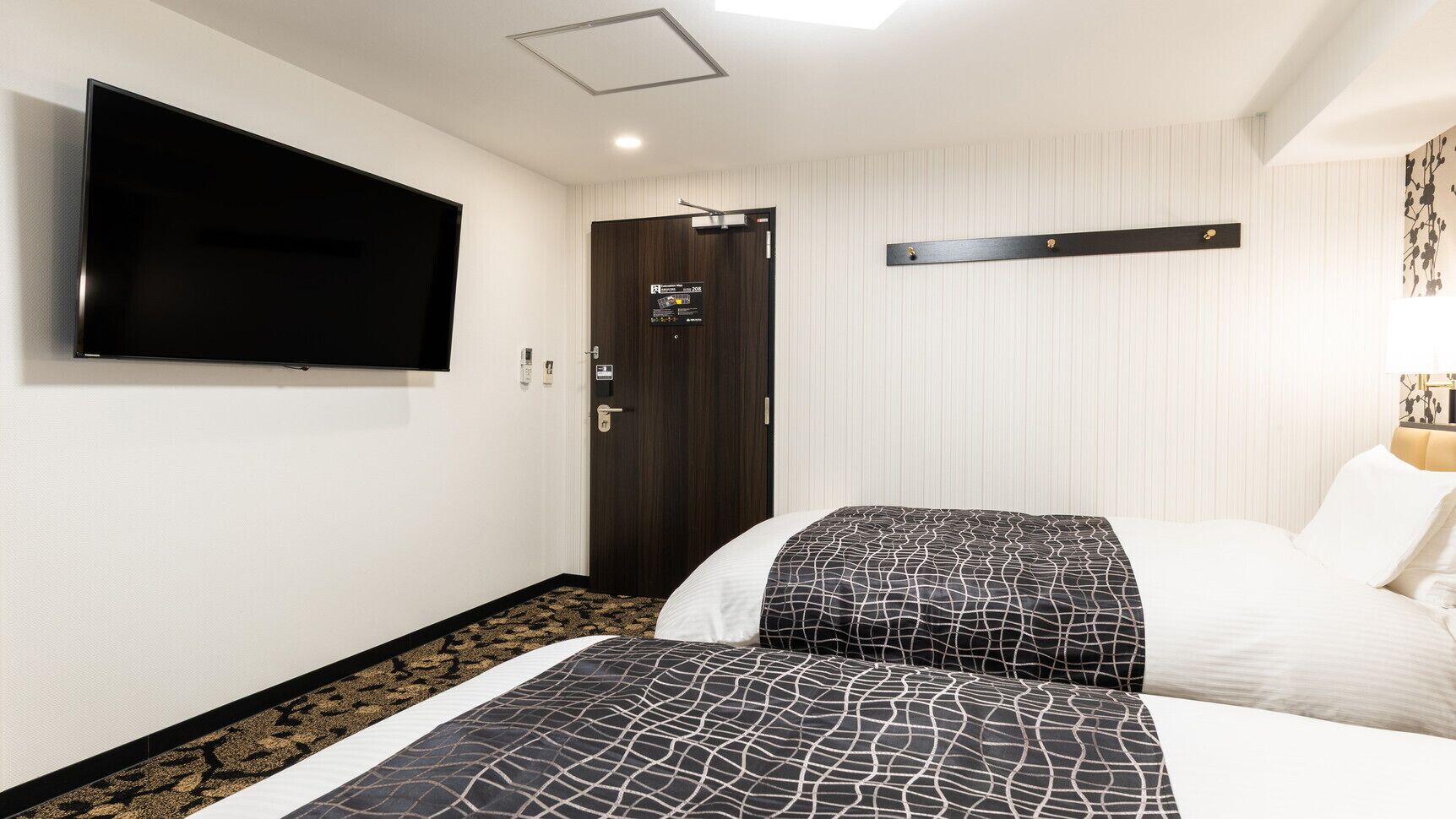 A hotel room with a bed, a TV mounted on the wall, a door, and a light fixture.