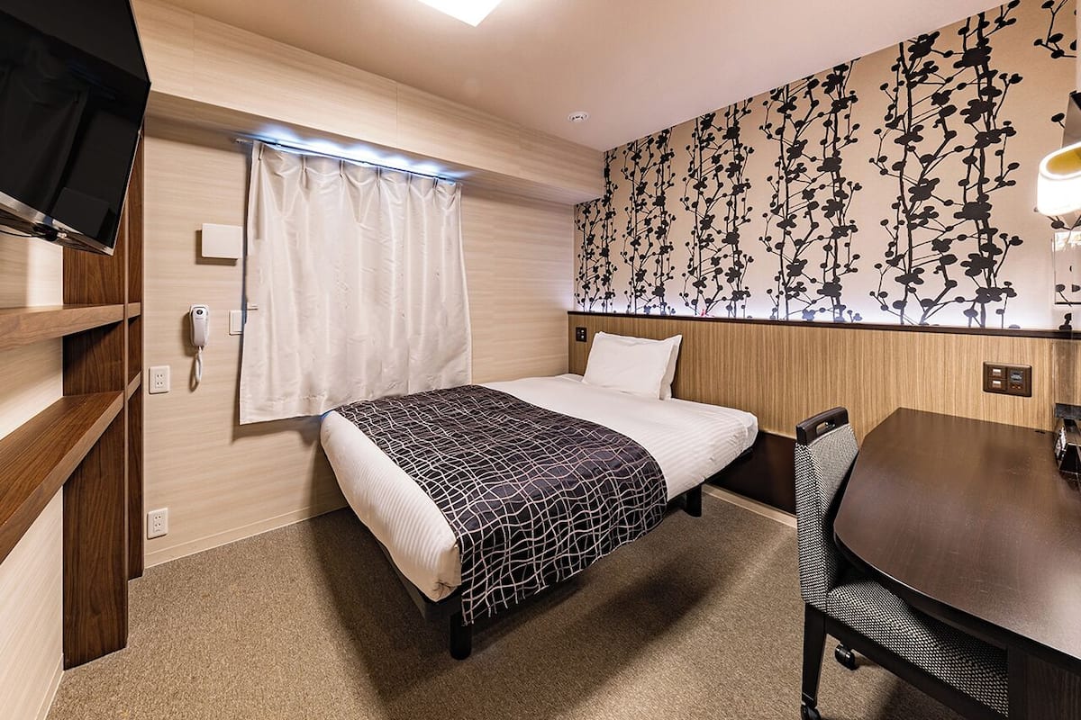 Ein Hotelzimmer mit Bett, Schreibtisch und Stuhl. Es gibt ein Fenster mit Vorhängen, einen an der Wand montierten Fernseher und eine Wand mit Blumenmuster.