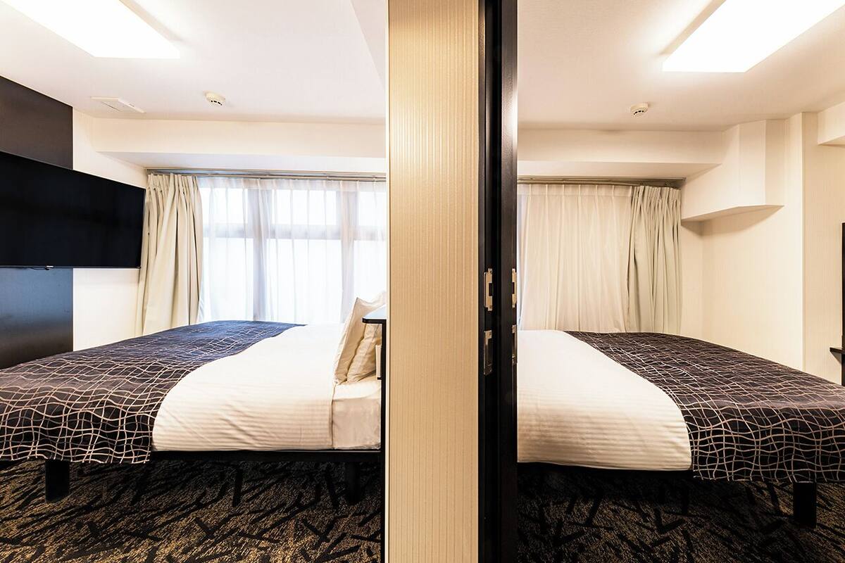 Ein Hotelzimmer mit zwei Betten, einem Flachbildfernseher, einem großen Fenster mit Vorhängen und einem Teppichboden.