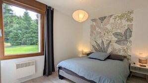 A bedroom with a bed, a window with curtains, a wall with a leaf pattern, a hanging light, and a bedside table with a lamp. - Quiet & Elegant Getaway: Modern Farmhouse with Terrace, Trails & No Overlook (Le Tholy)