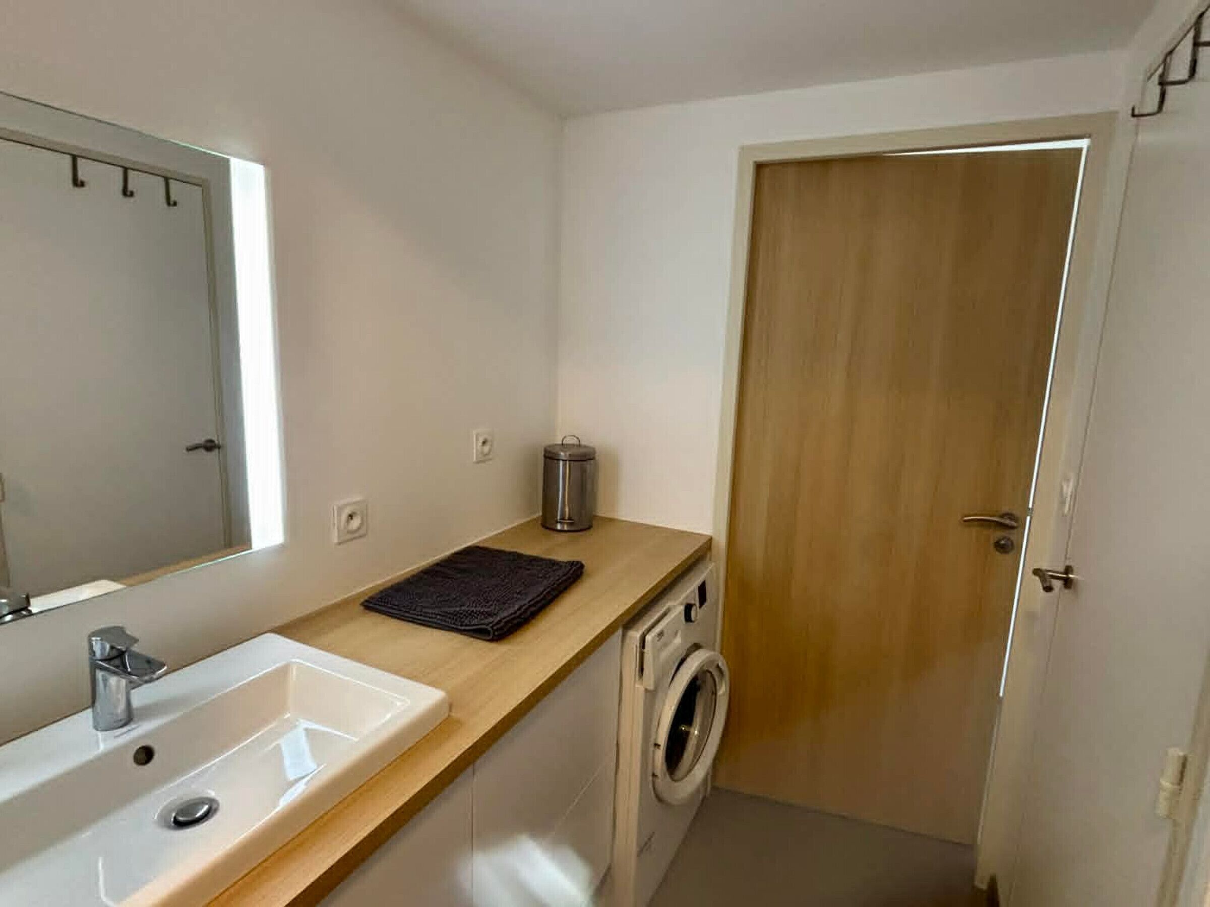 A compact bathroom with a sink, mirror, washing machine, and a small trash bin.