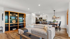 A modern living room with a sectional sofa, a wooden bookshelf, a dining area with a table and chairs, and a kitchen in the background. - Hauts-Bois Ski In/Out 2brs w/Pool Access (193-1) (Mont-Tremblant)