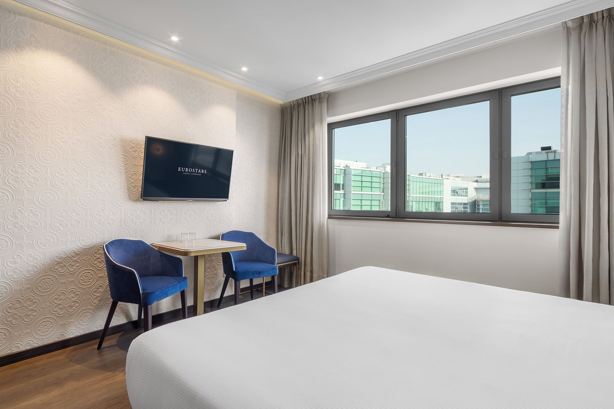 Habitación de hotel con cama, dos sillas azules, una mesita, televisor en la pared y un ventanal con vista a la ciudad.