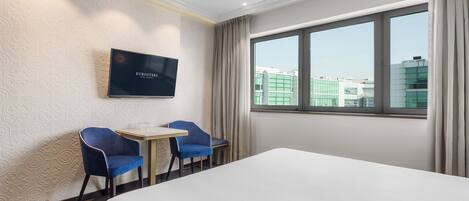 Habitación de hotel con cama, dos sillas azules, una mesita, televisor en la pared y un ventanal con vista a la ciudad.