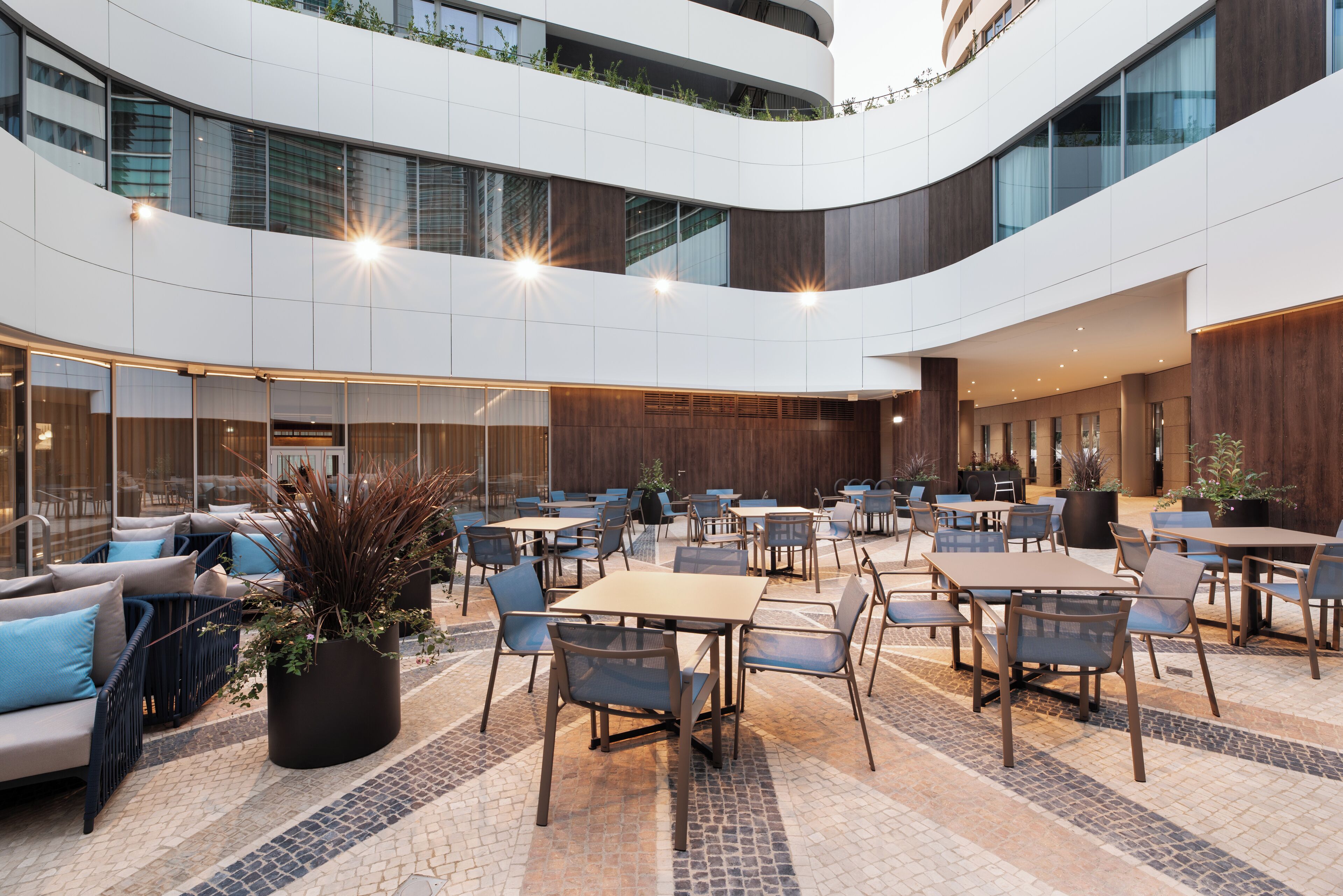 A spacious indoor courtyard with multiple seating areas, including tables and chairs, surrounded by large windows and a modern architectural design.