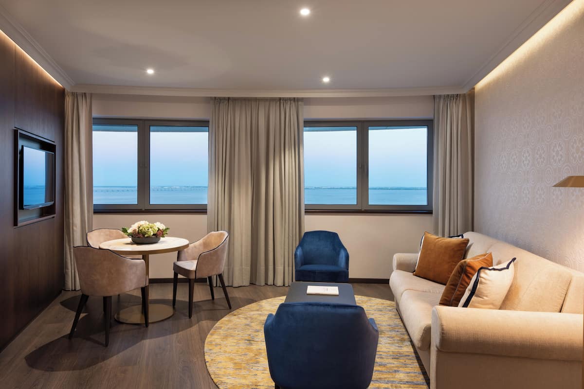 Ein modernes Hotelzimmer mit einem runden Esstisch, zwei Stühlen, einer Couch und einem Fernseher. Große Fenster bieten Blick auf das Meer.