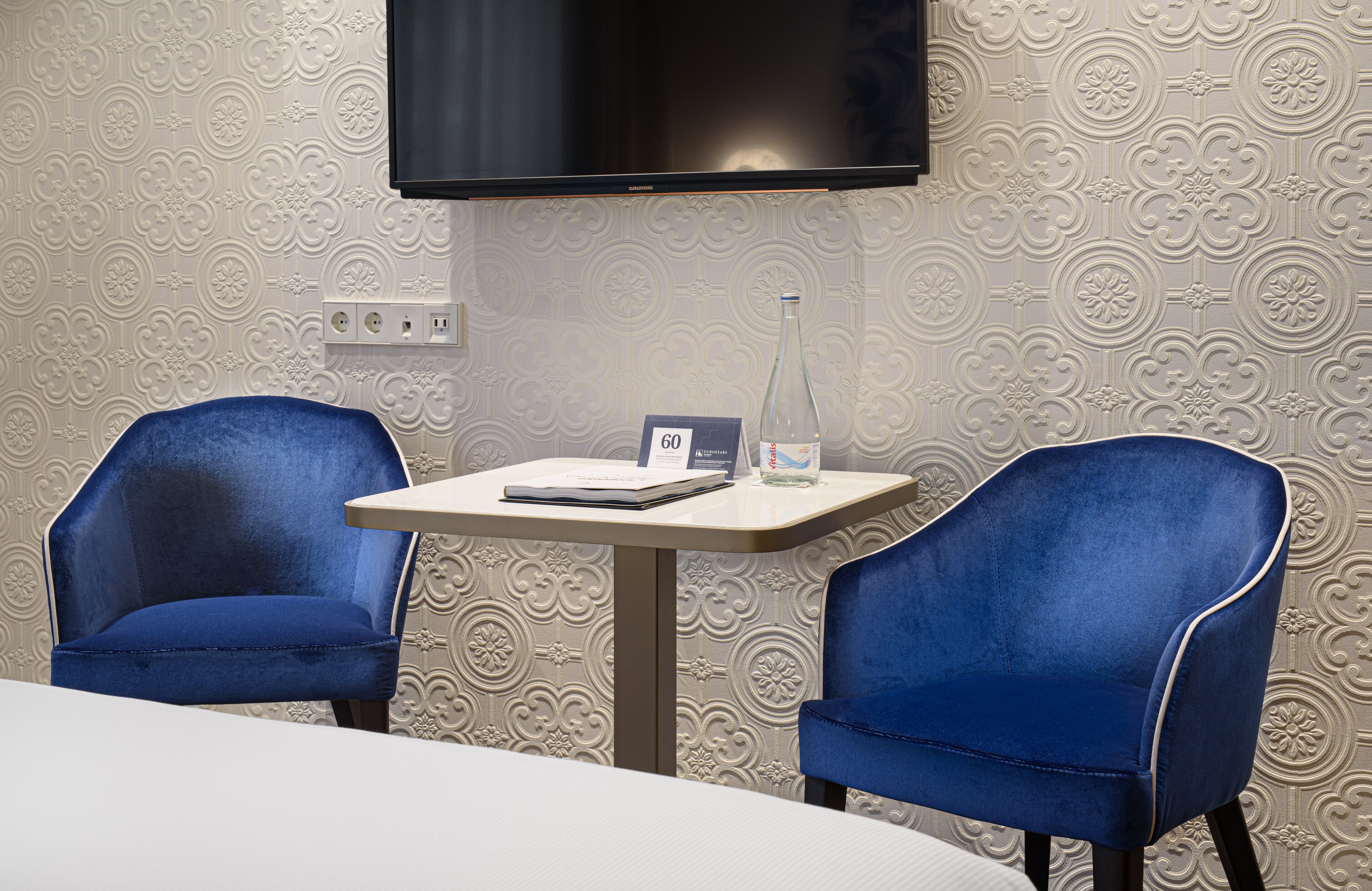 A hotel room with two blue armchairs, a small table with a bottle and a book, and a TV mounted on the wall.