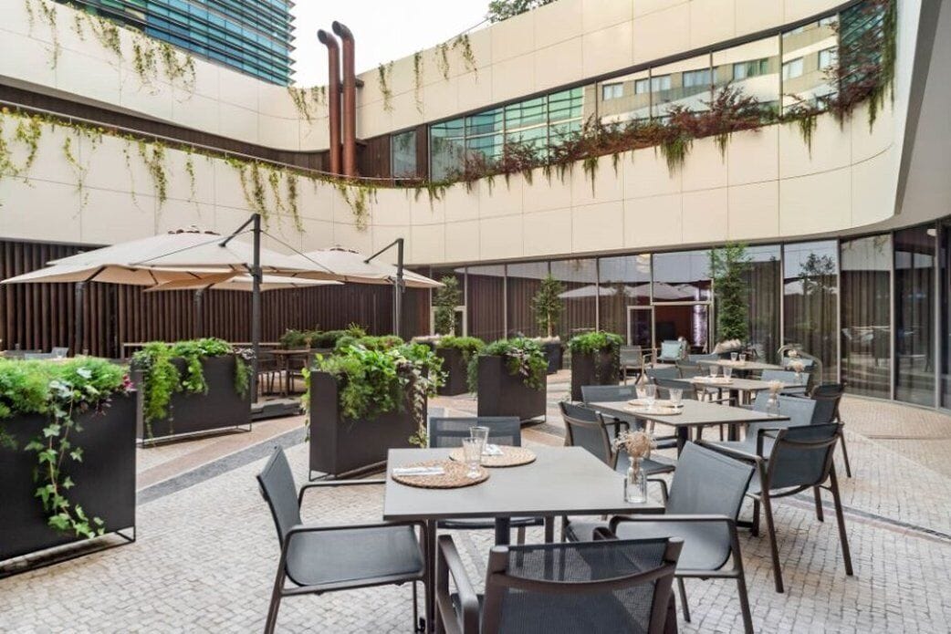 Un espace extérieur aménagé pour les repas, avec des tables et des chaises, entouré de jardinières remplies de verdure, et un bâtiment moderne aux grandes fenêtres en arrière-plan.
