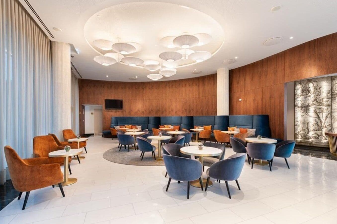 A modern hotel lobby with round tables, blue and orange chairs, a wooden wall, and a large decorative light fixture.