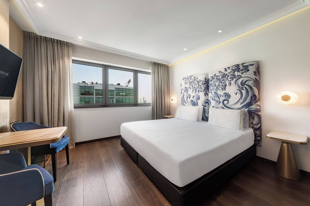Ein Hotelzimmer mit einem großen Bett, einem Schreibtisch, einem Stuhl, einem Fenster mit Vorhängen und einem an der Wand hängenden Kunstwerk.