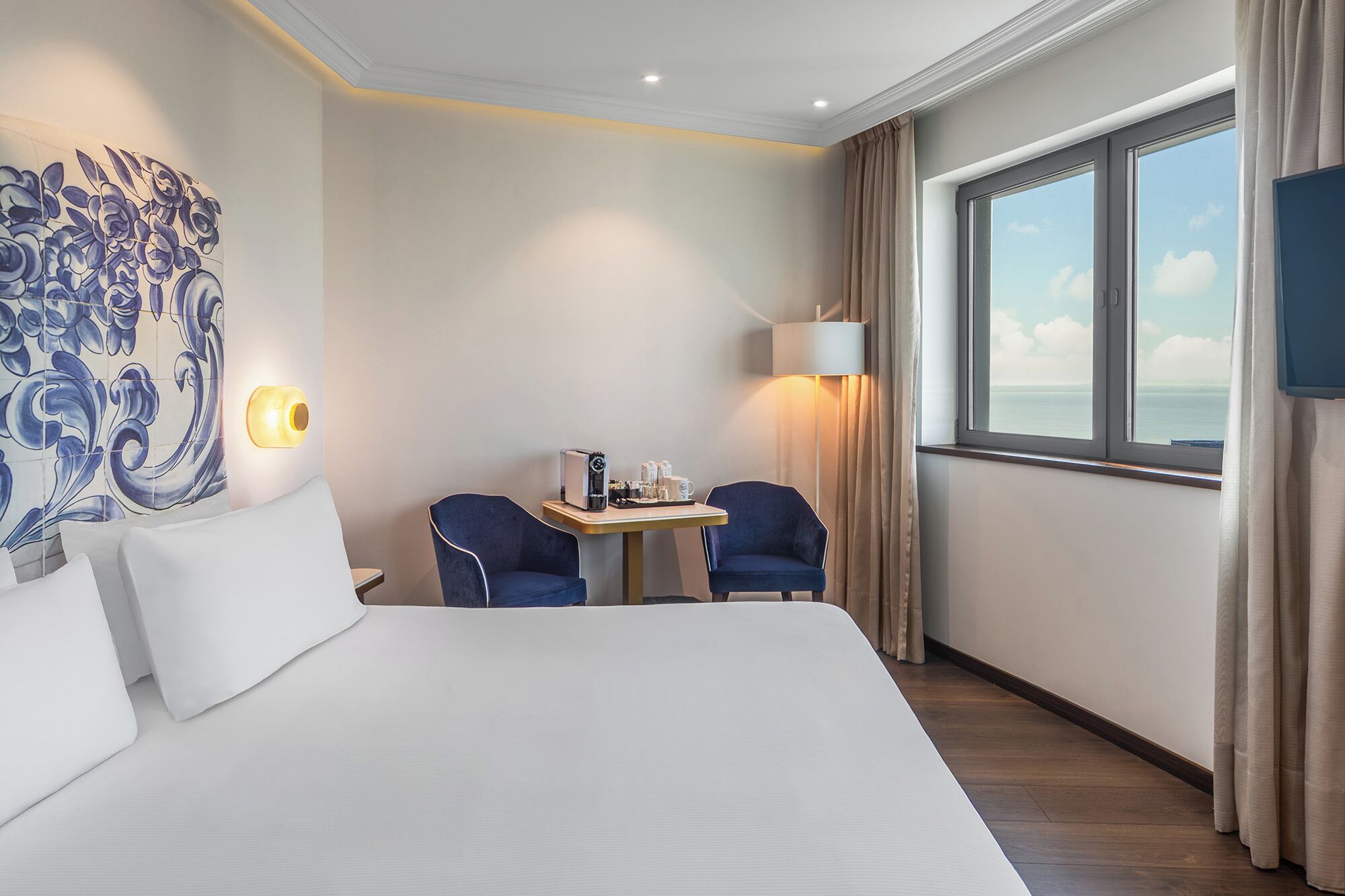 A hotel room with a large bed, two chairs, a small table with a coffee maker, a lamp, and a TV.