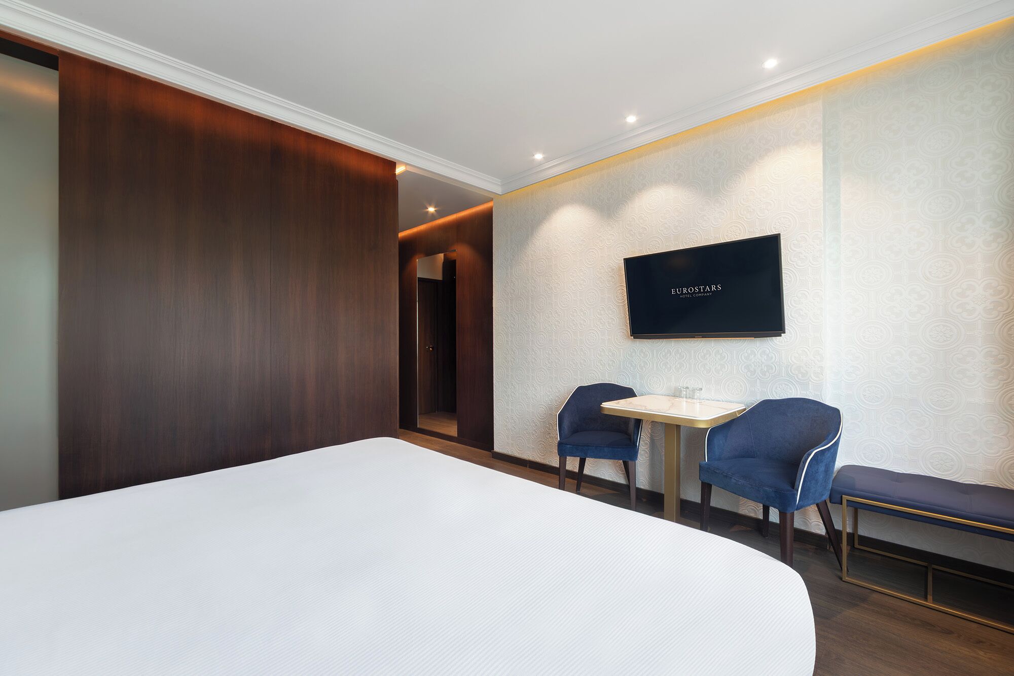Ein Hotelzimmer mit einem großen Bett, zwei blauen Sesseln, einem kleinen Tisch und einem Fernseher an der Wand.