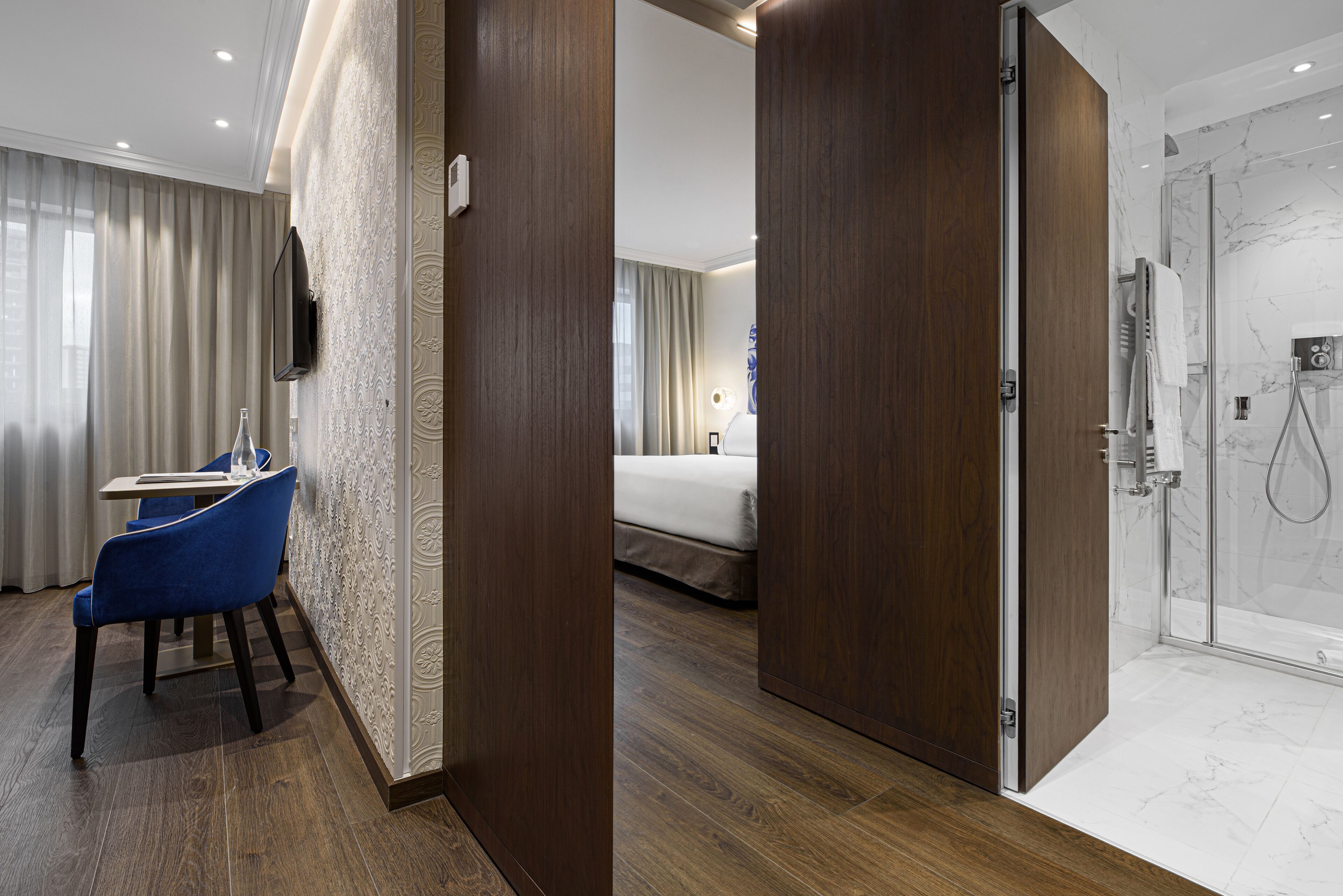 Une chambre d’hôtel avec un plancher en bois, un lit, une salle de bain avec douche et un coin salon comprenant une table et des chaises.