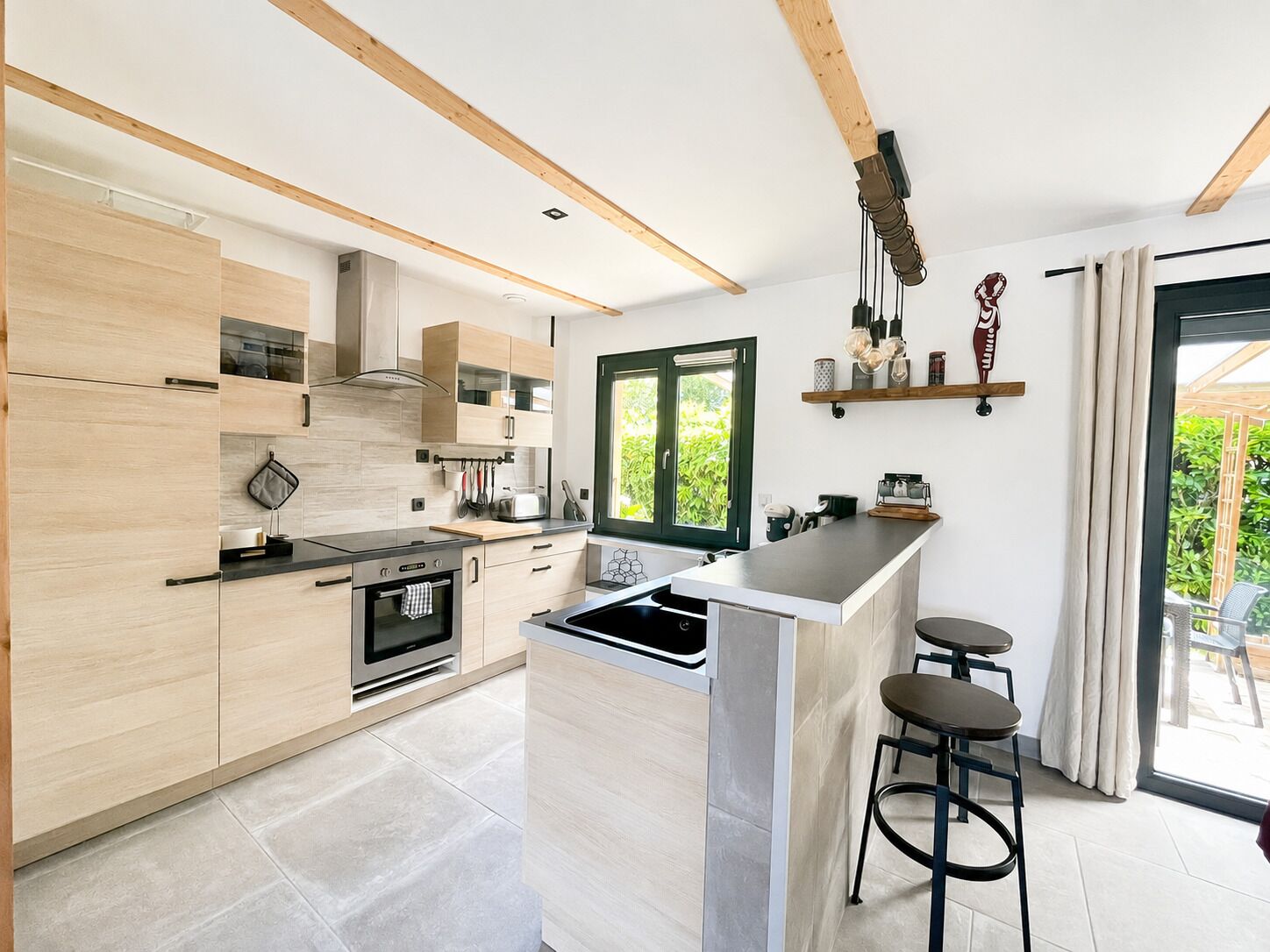 A modern kitchen with wooden cabinets, a built-in oven, a large island with bar stools, and a window providing a view of the outdoors.
