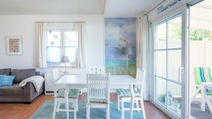 Een lichte woonkamer met een witte bank, een eettafel met stoelen, en een grote schuifpui die leidt naar een buitenruimte met een groene stoel.