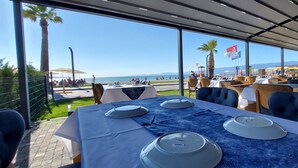 Un área de comedor al aire libre con mesas dispuestas para el servicio, con vista a una playa con palmeras y cielo despejado.
