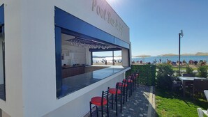 Un bar y cafetería junto a la piscina, con mostrador de bar y taburetes rojos, con vista a una playa con sombrillas y gente.