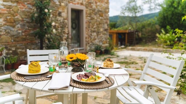 An outdoor dining table set with plates of food, surrounded by white chairs, in front of a stone building with a door and a garden. - Le Fonti - Sasso: large and bright (Pontassieve)