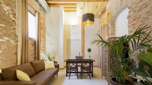 Una habitación acogedora con un sofá de cuero marrón, una mesa de comedor de madera con sillas y una computadora portátil. Hay plantas en macetas y un separador decorativo con una planta al lado derecho.
