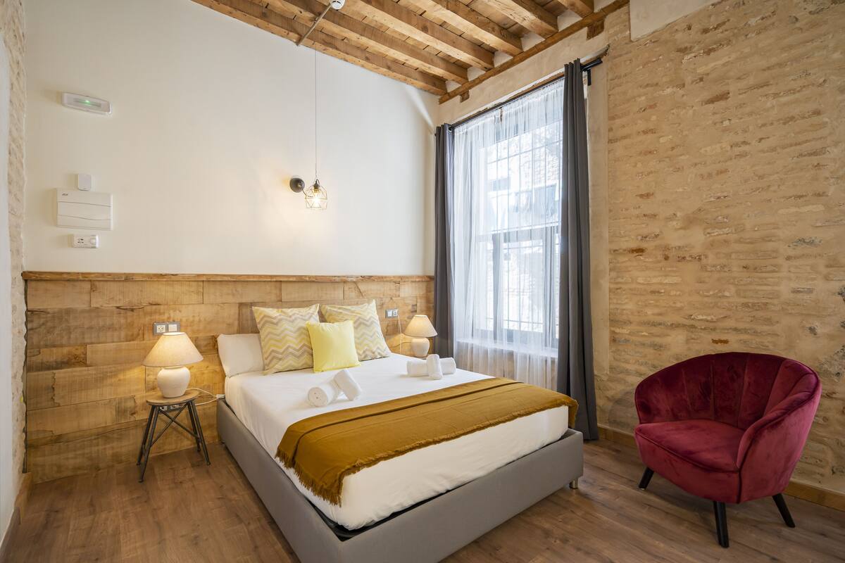Un dormitorio con cabecera de madera, cama con ropa de cama blanca, una silla roja y un ventanal con cortinas traslúcidas.