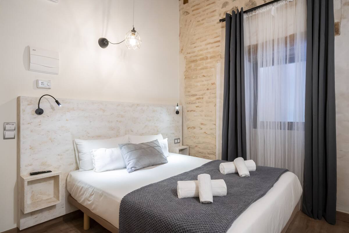 Una cama bien tendida con una manta gris y toallas enrolladas, un cabecero con una lámpara, una mesita de noche con un control remoto y una ventana con cortinas traslúcidas.