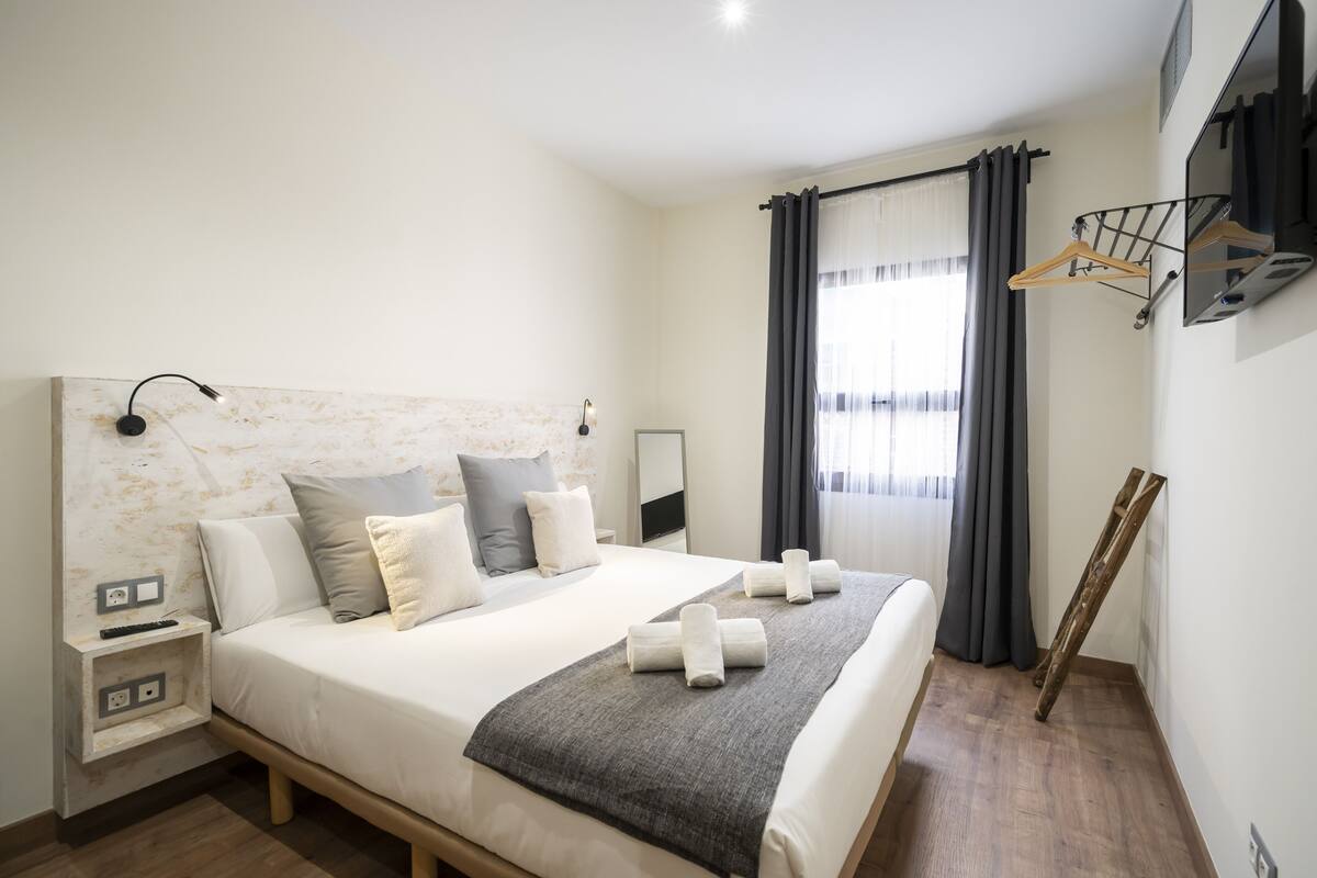 Una habitación de hotel moderna con una cama grande, mesitas de noche, un escritorio, un espejo y una ventana con cortinas.
