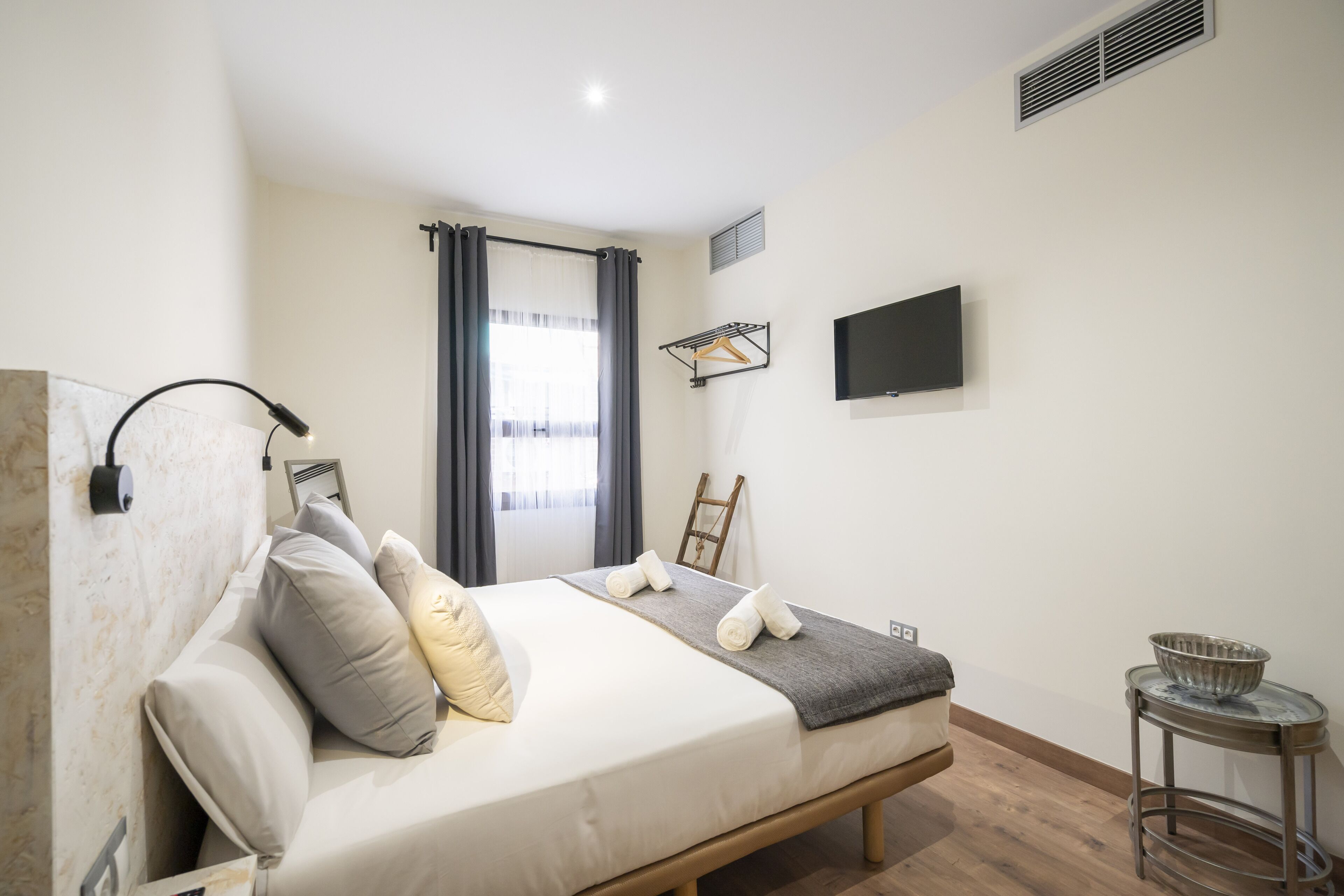 A bedroom with a bed, a bedside table, a TV mounted on the wall, and a window with curtains.